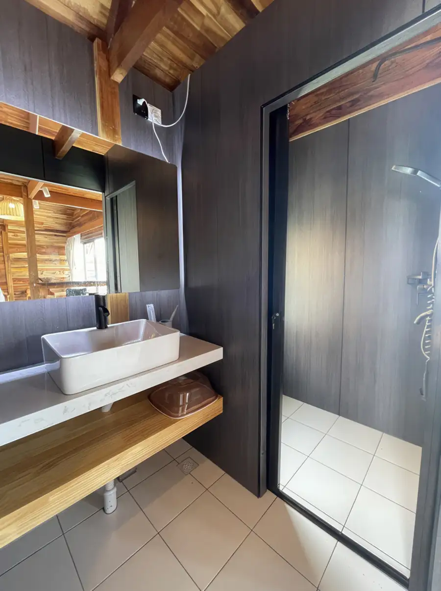 An elegant bathroom vanity area at Jelly Sea Resort highlighting a sleek light wood desk, a glowing backlit round mirror, a white ceramic washbasin, and an electric shower.