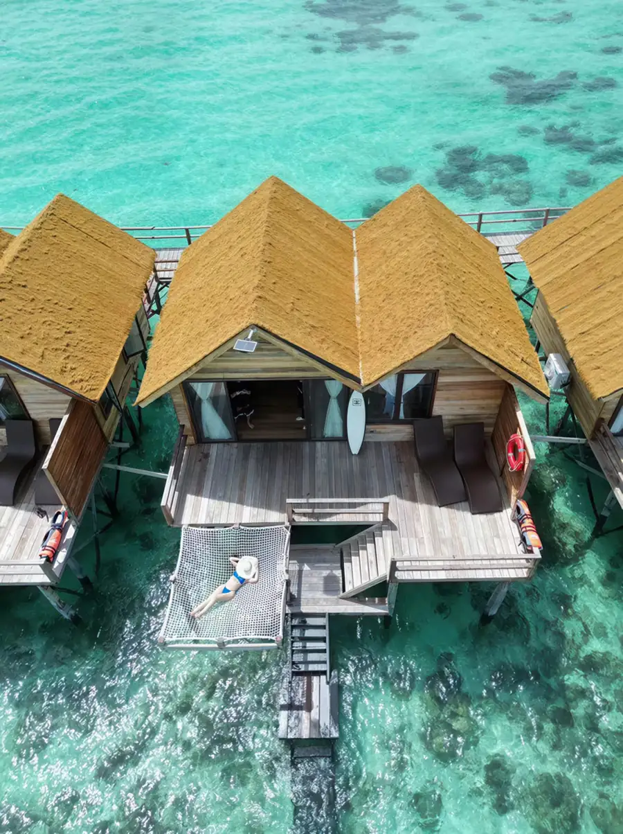 A low-level aerial view skimming past white overwater chalets that highlight private balconies and overwater hammocks, reflecting native maritime charm at Jelly Sea Resort.