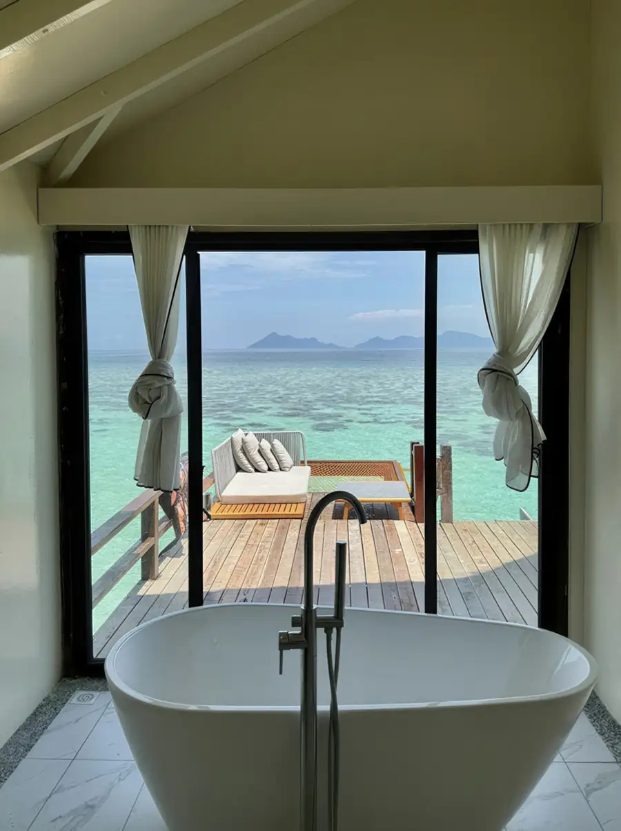 A luxurious white bathtub with a floor-mounted faucet looking out onto a private wooden deck with lounge seating and clear turquoise waters at Jelly Sea Resort.