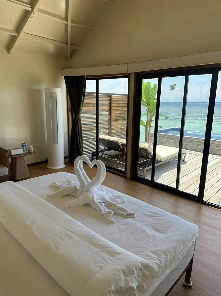 A relaxing bedroom featuring a bed decorated with towel swans, a floor-standing A/C unit, and sliding glass doors that lead to a balcony with a stunning seaview over Tun Sakaran Marine Park.