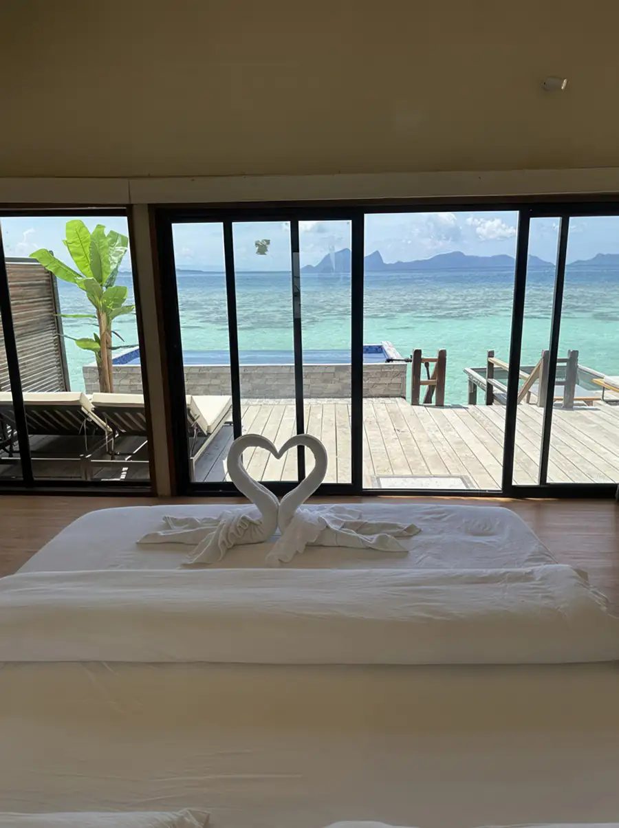 A beautiful bedroom at Jelly Sea Resort near Semporna highlighting a bed with swan towel art, large glass windows with seaviews, warm wooden plank floor with varnished timber, and aircon.