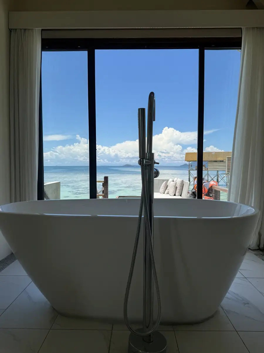 A sleek white freestanding bathtub featuring a floor-mounted chrome hand shower with a direct view of the ocean and nearby water villas at Jelly Sea Resort.